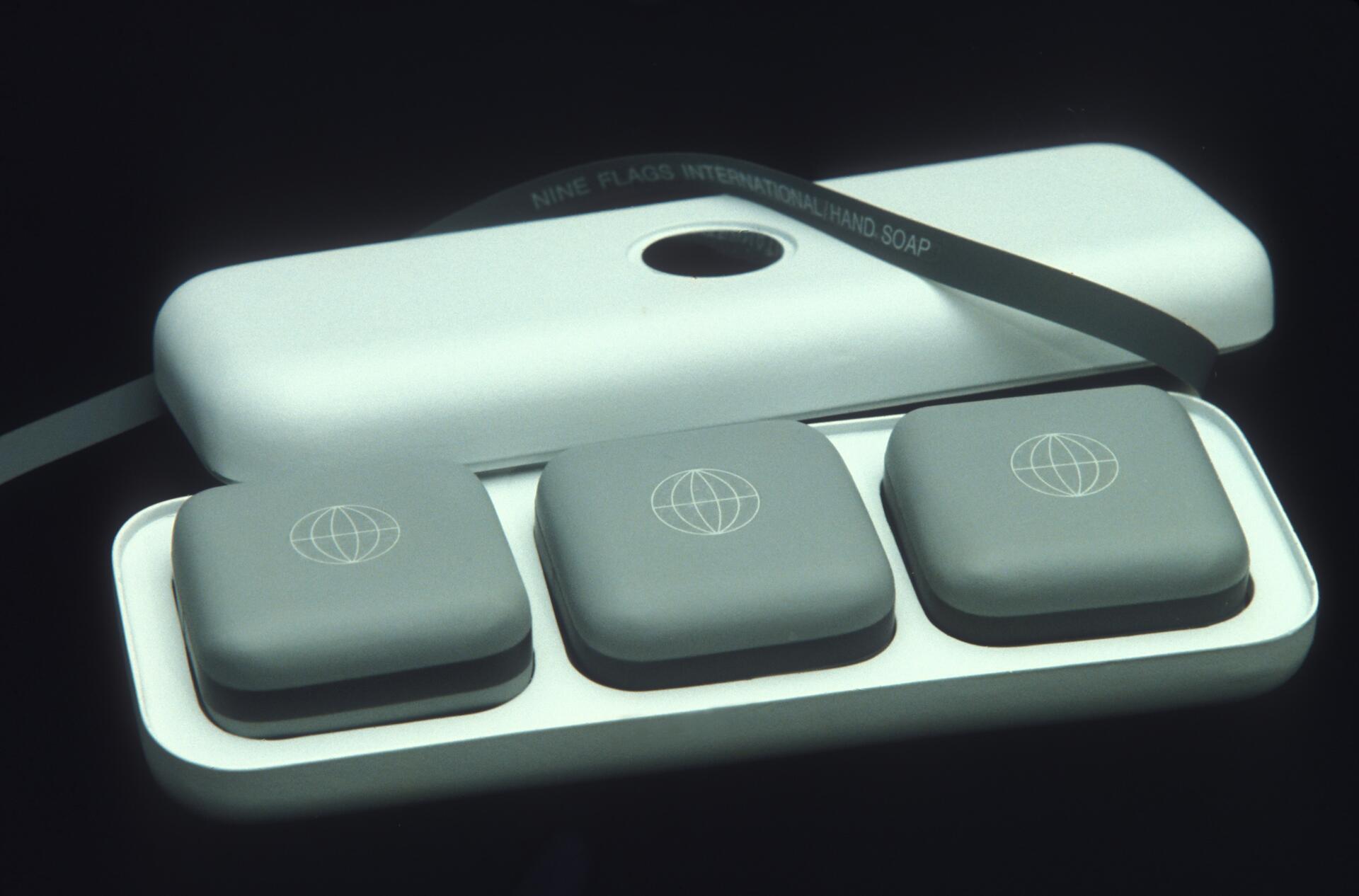 A white soap case with three gray bars of soap, each featuring a simple globe logo. A ribbon reads "Nine Flags International Hand Soap."