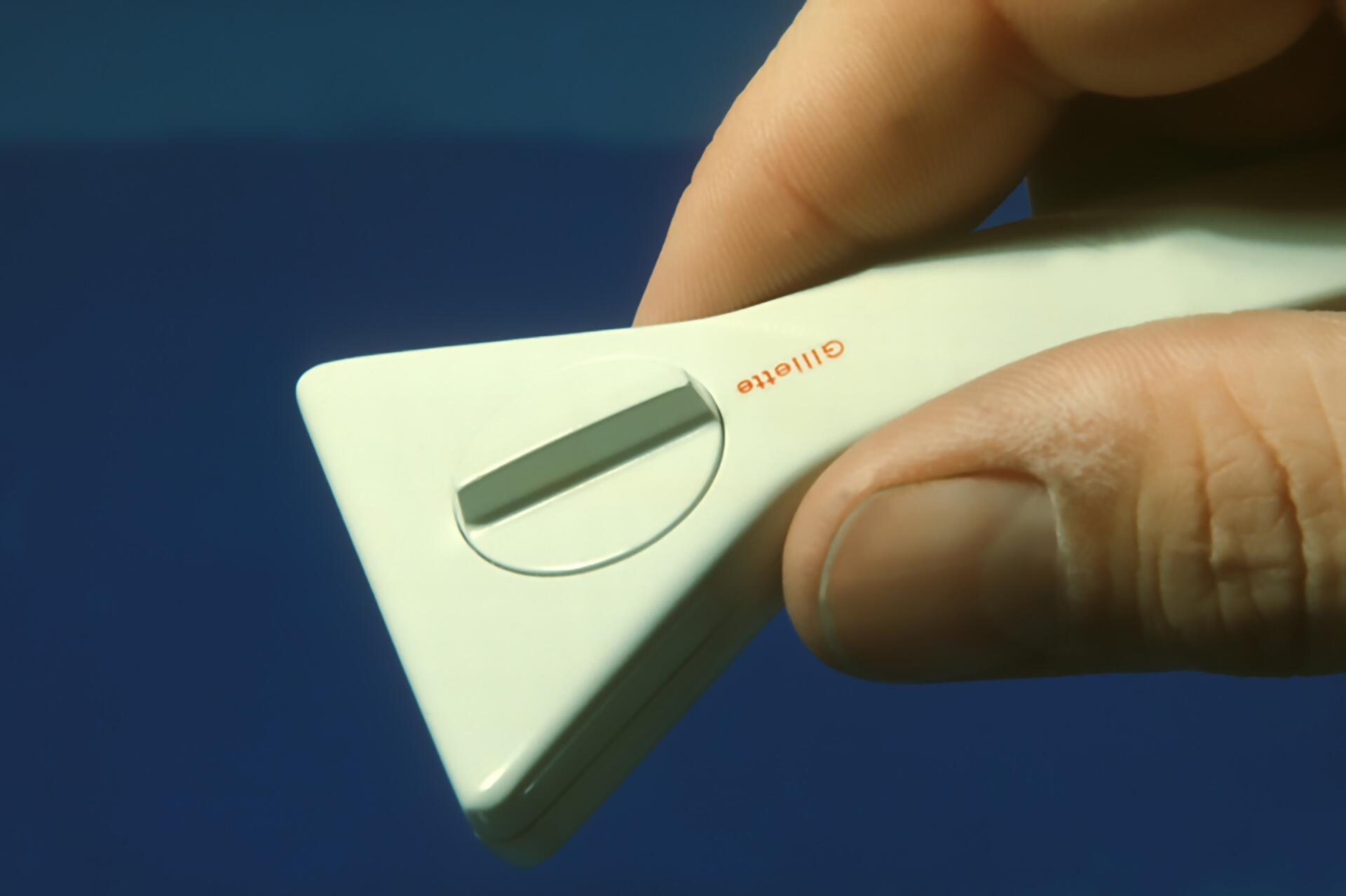 Hand holding a white Gillette razor with a triangular shape and circular blade cover, set against a blue background.