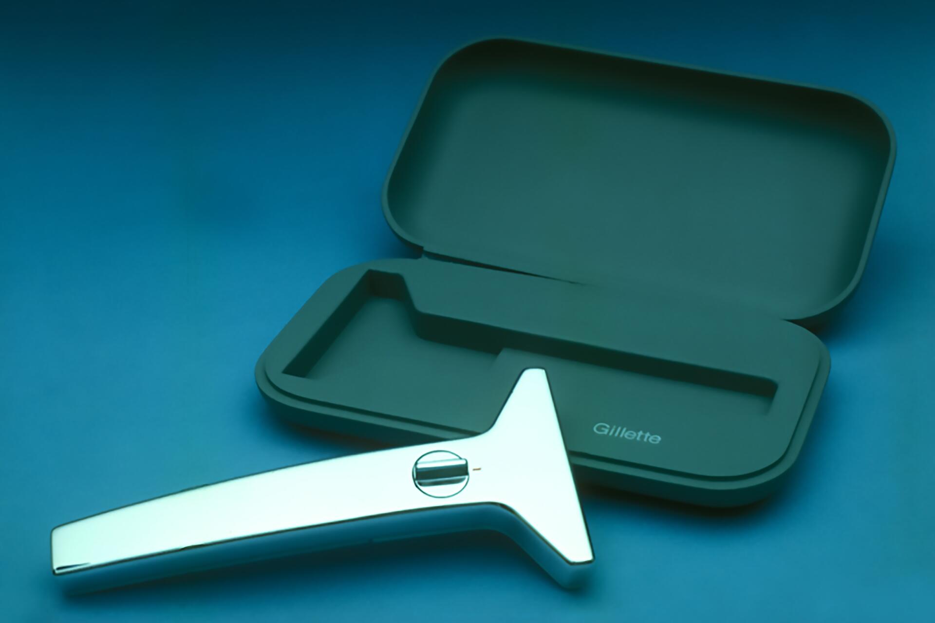 Sleek silver razor with a T-shaped handle next to an open dark gray case with the brand name embossed inside, on a teal background.