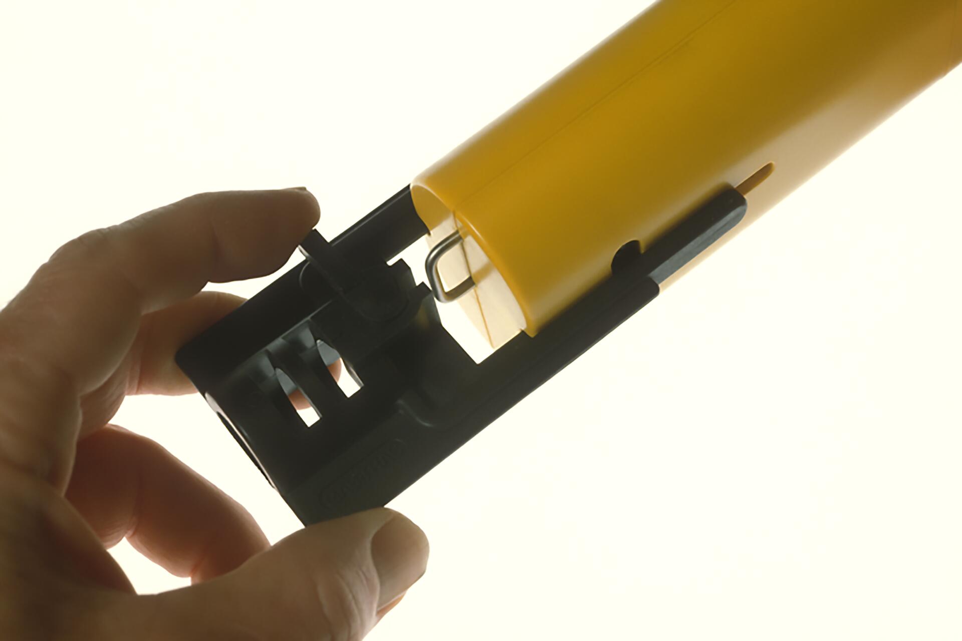 A hand holding a black and yellow plastic device, revealing its internal mechanism with a visible wire loop, set against a light background.