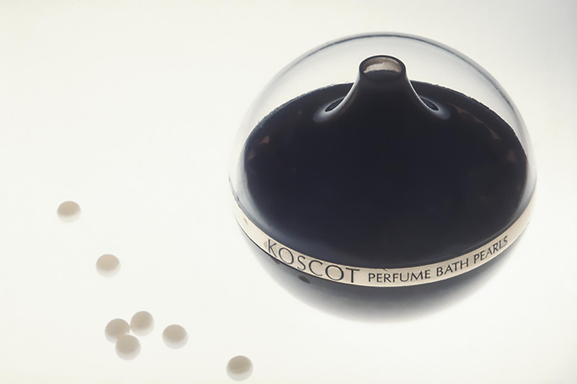 Clear dome-shaped container labeled "Koscot Perfume Bath Pearls" with light-colored pearls scattered nearby on a white surface.