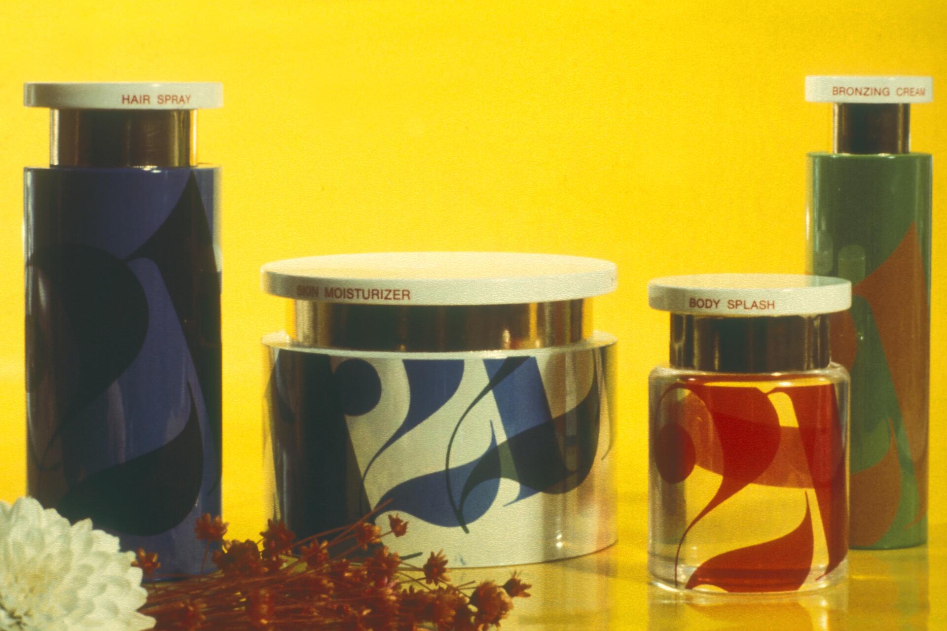 Four decorative cosmetic containers with labels for hair spray, skin moisturizer, body splash, and bronzing cream, set against a bright yellow background. White flowers and small dried plants are in the foreground.