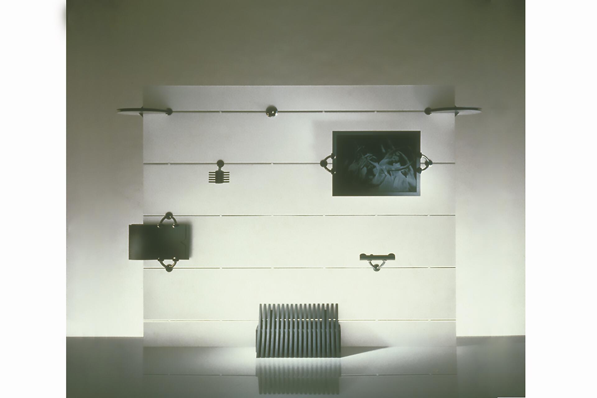 Minimalist wall installation with horizontal lines and mounted black objects, including abstract designs and a photograph. Dark geometric shapes, reflection on shiny surface below, beige background.