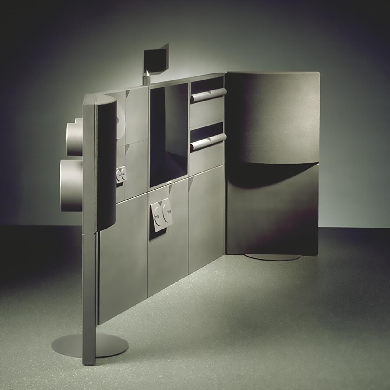 Futuristic modular wall with geometric shapes, metallic panels, and cylindrical protrusions, standing on a speckled floor in a dimly lit room.