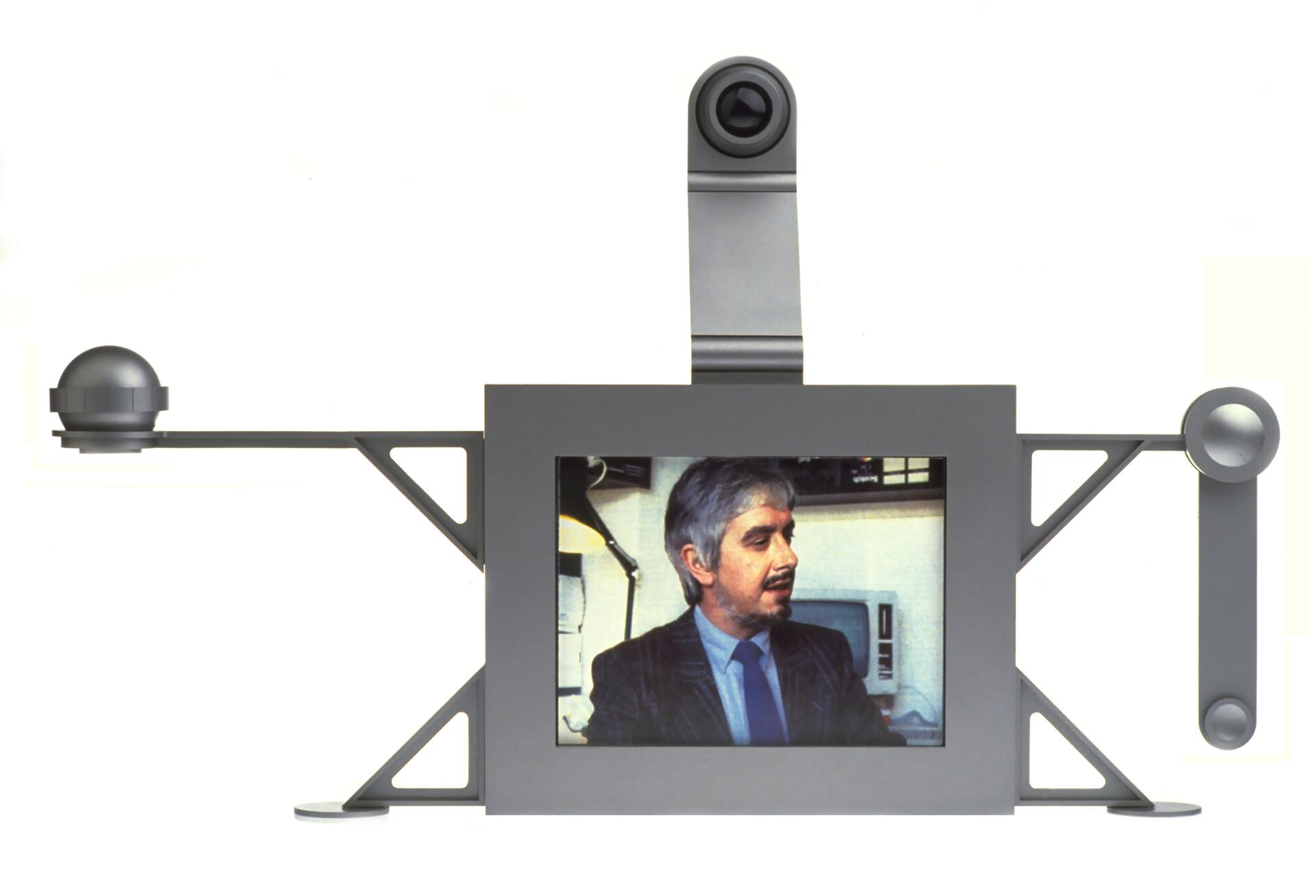 Futuristic video conferencing device with a central screen showing a man in a suit, flanked by angular supports and round sensors on both sides, topped by a camera.