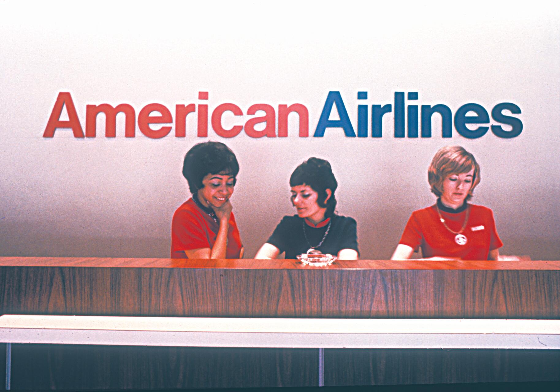 Three women wearing red uniforms sit behind a wooden counter under the American Airlines logo.