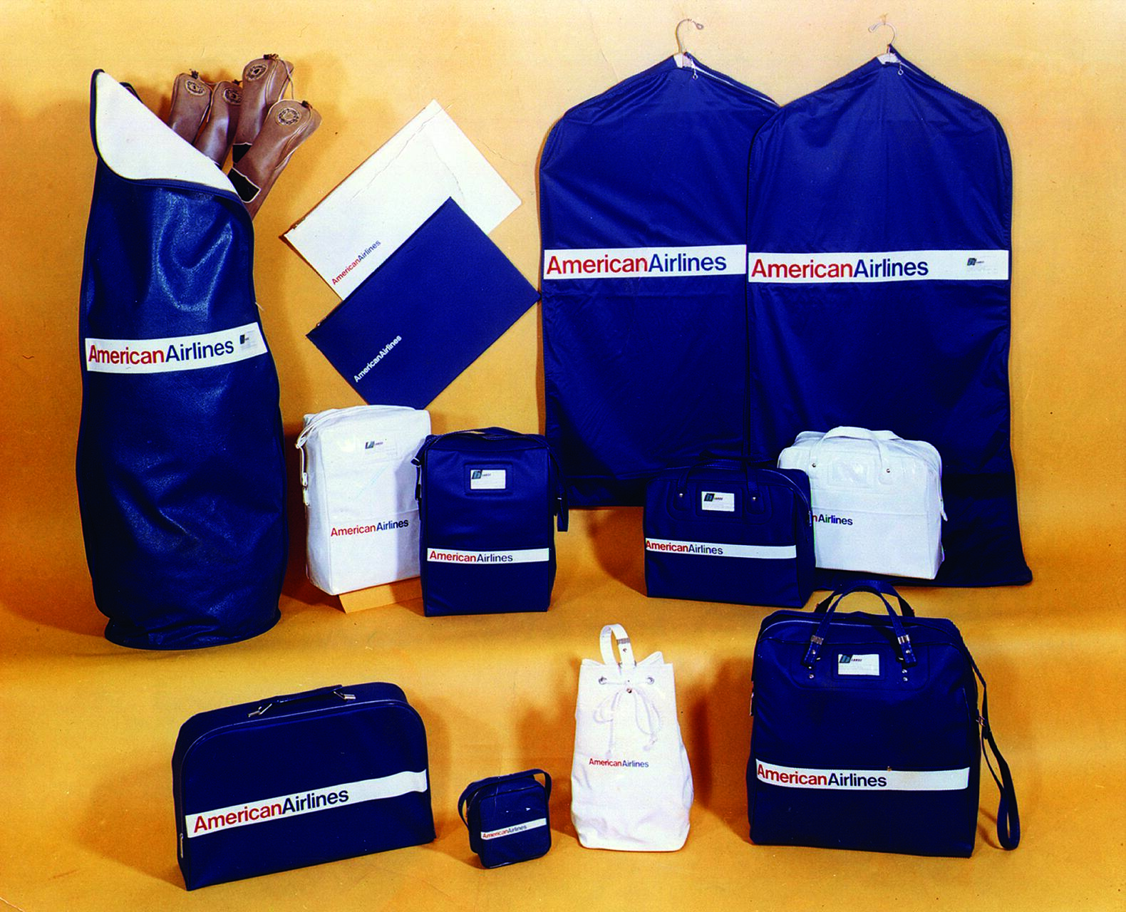Various blue and white American Airlines branded bags and travel accessories arranged on a beige background, including garment bags, duffel bags, and small pouches.
