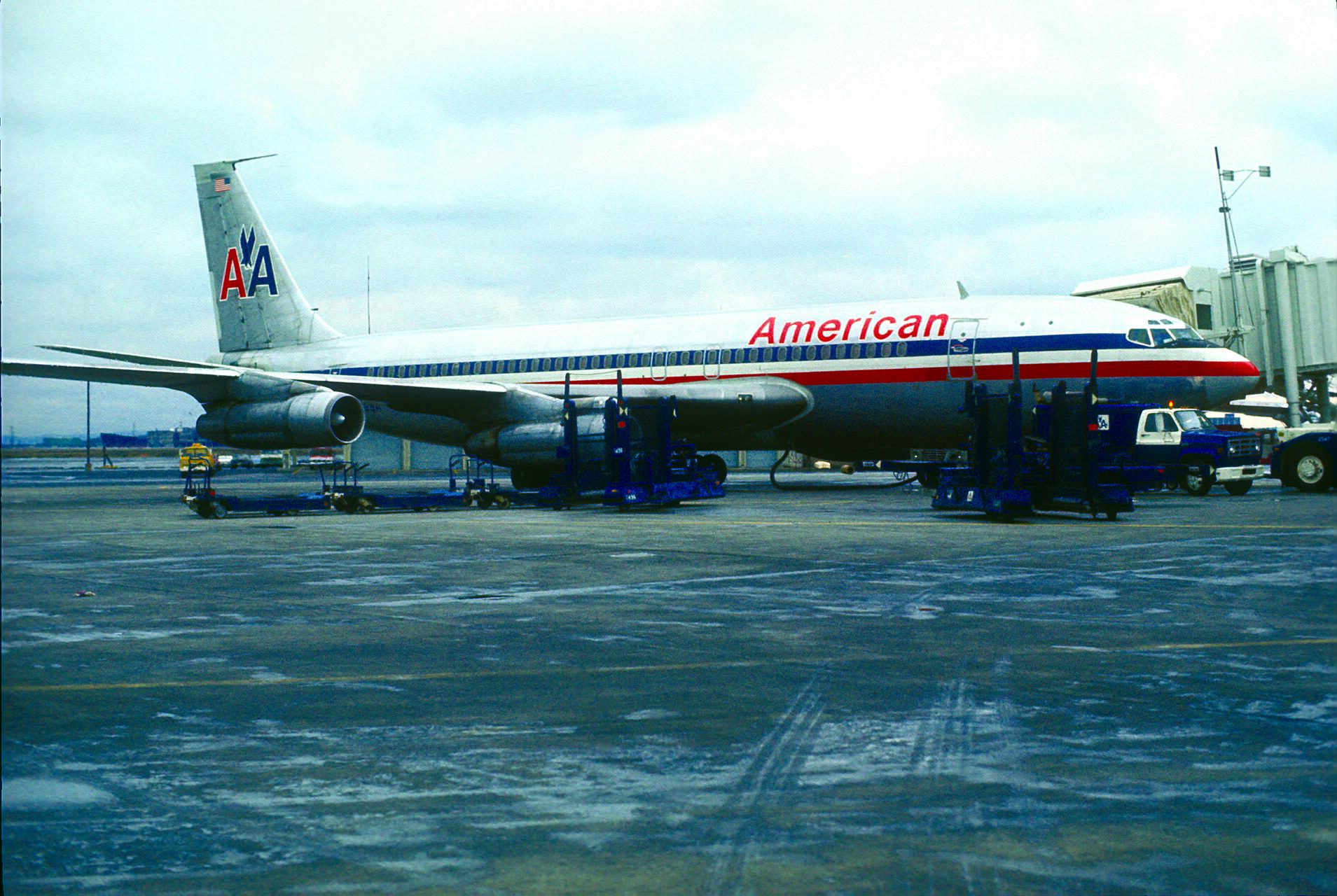 Aircraft at an airport gate with an American Airlines livery, featuring red, white, and blue stripes. Ground crew equipment and vehicles are nearby under a cloudy sky.
