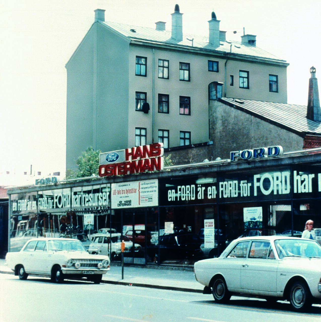 Street view of an old Ford dealership with large signage reading "Hans Osterman." Two vintage cars are parked in front. A multi-story building with several chimneys is visible in the background.
