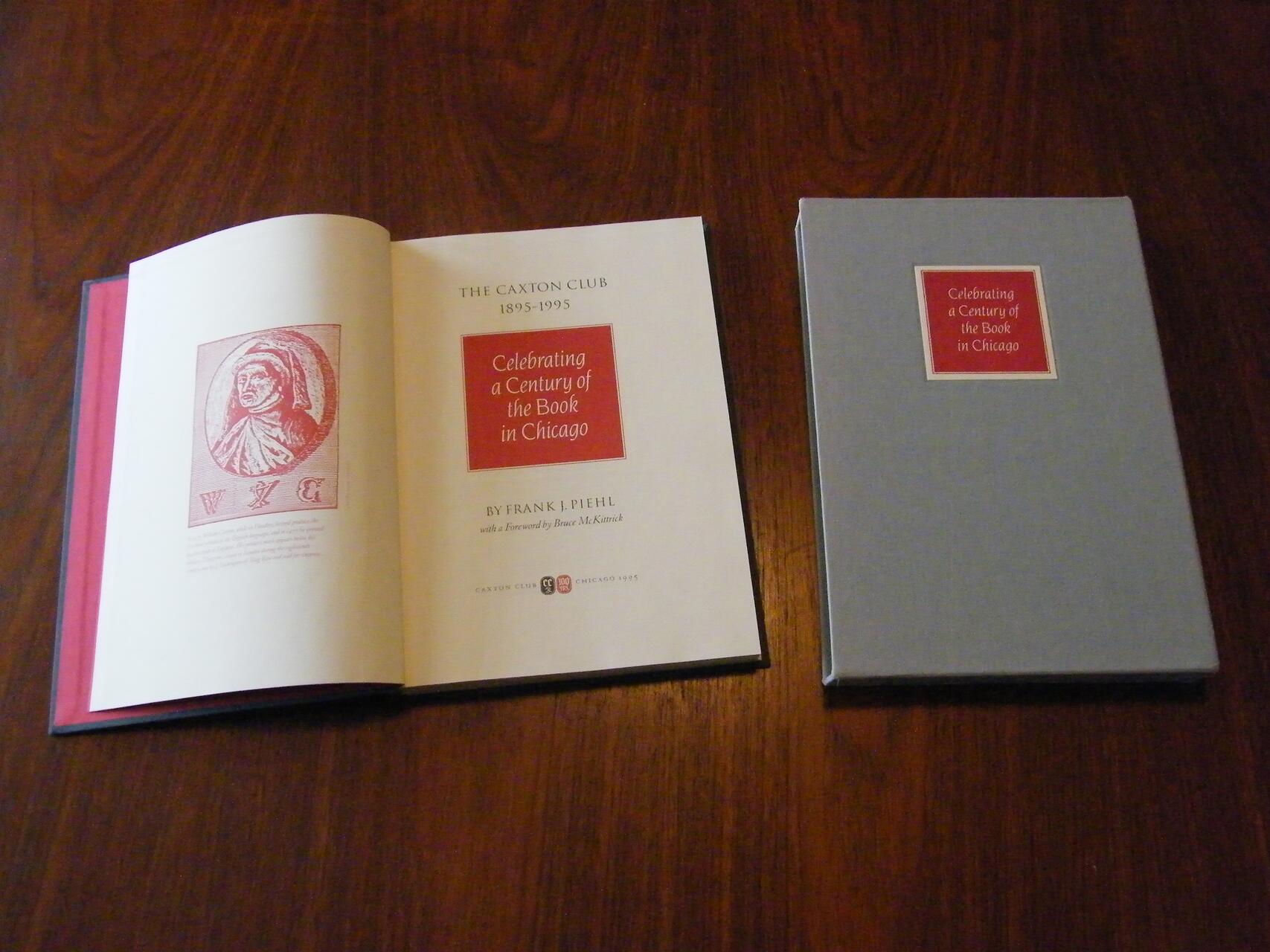 Open book with title "Celebrating a Century of the Book in Chicago" next to a closed blue hardcover with the same red label on a wooden surface.