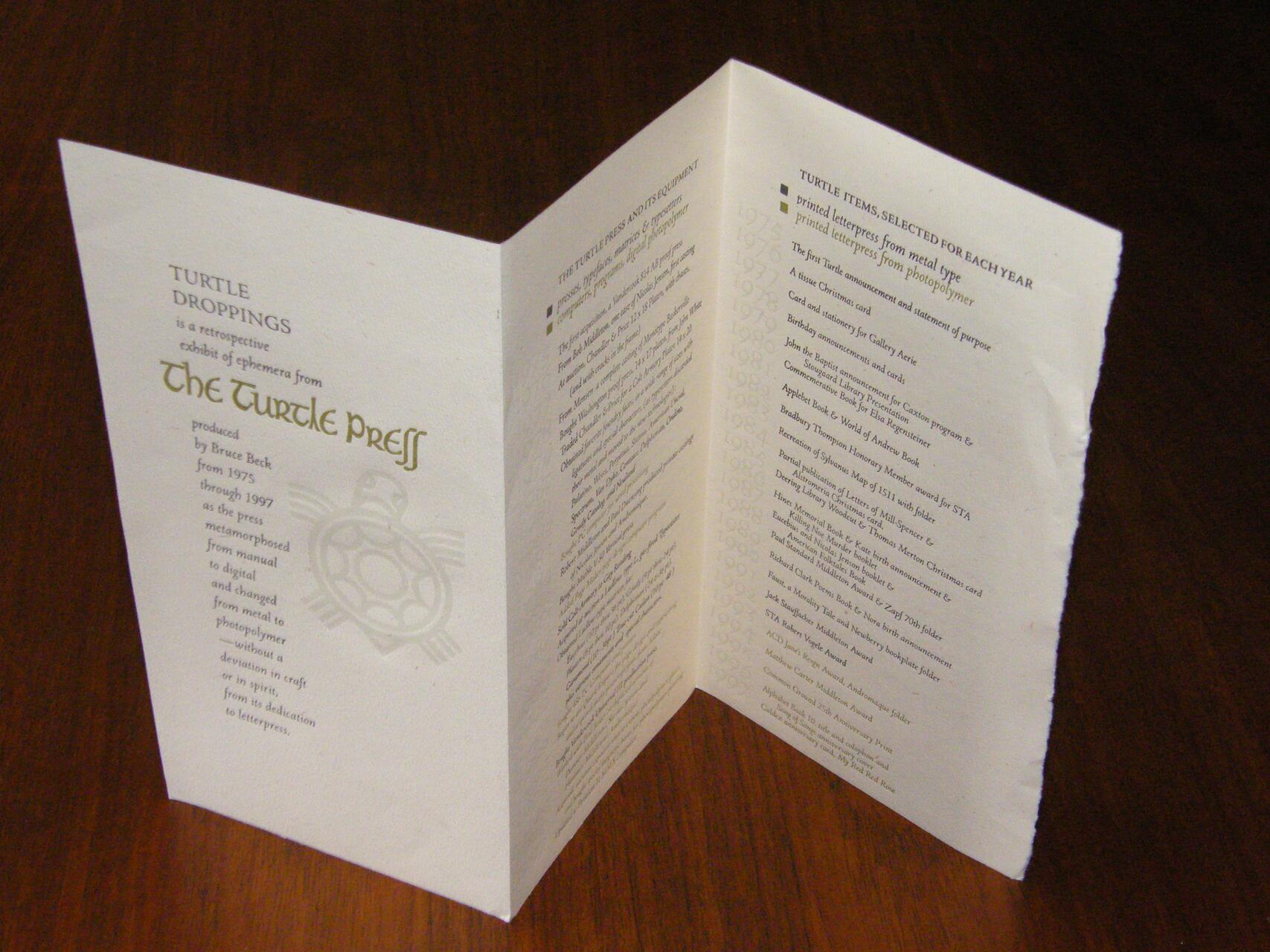 Tri-fold brochure on a wooden table with text about "The Turtle Press" and a turtle graphic. The inside lists selected items from various years in two columns. The paper has a rustic, textured appearance.