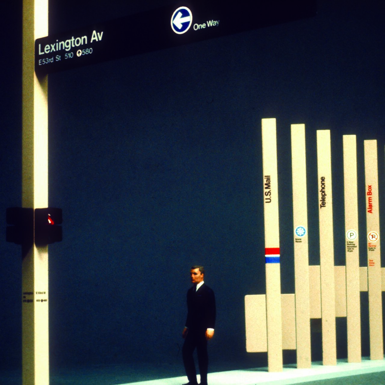 A man in a suit walks past a series of tall, vertical signs including U.S. Mail, Telephone, and Alarm Box. A traffic light shows red. The street sign reads "Lexington Av" with directional arrows. The background is dark, enhancing the illuminated signs.