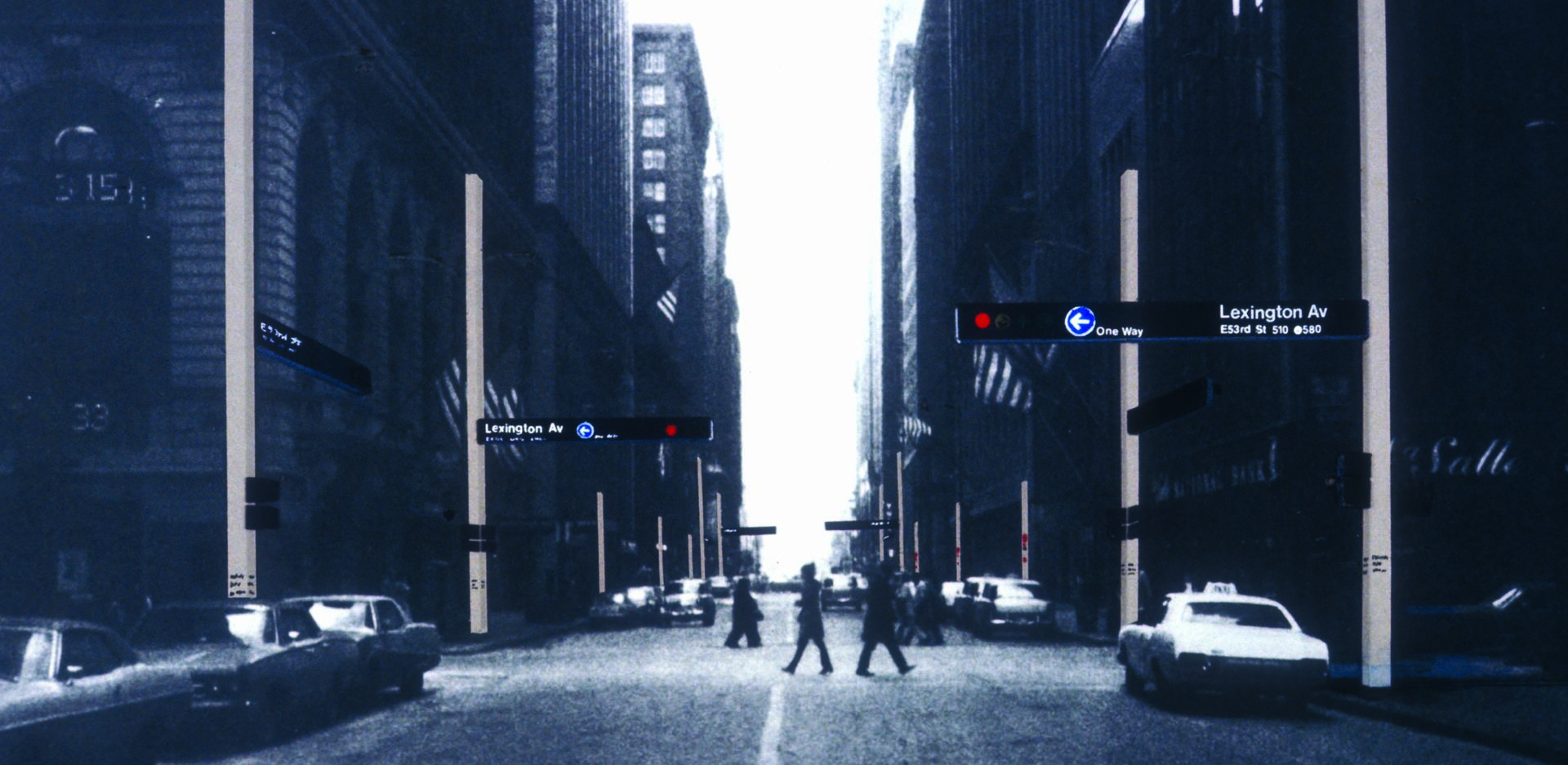A black-and-white photograph shows a city intersection with vintage cars and people crossing the street. Tall buildings line the background, with a one-way street sign and tall poles featuring directional signage like "Lexington Av".