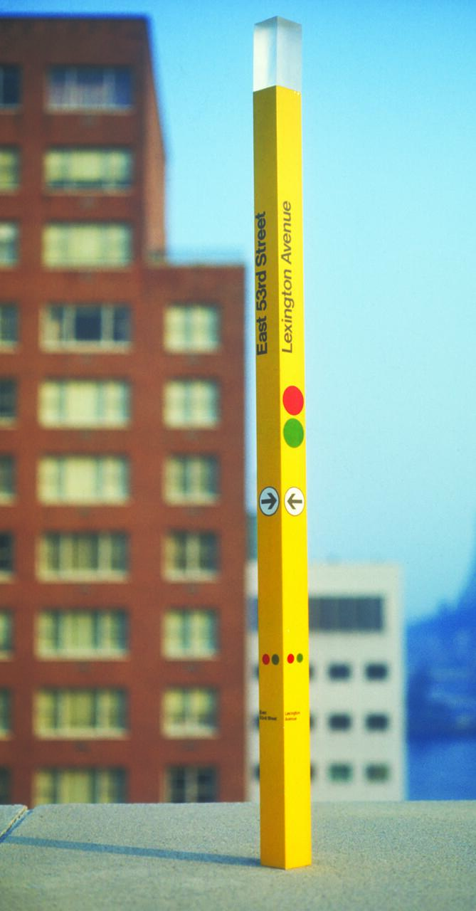 Tall, slender yellow signpost against a blurred cityscape, featuring circles and arrows with text reading "East 53rd Street, Lexington Avenue."