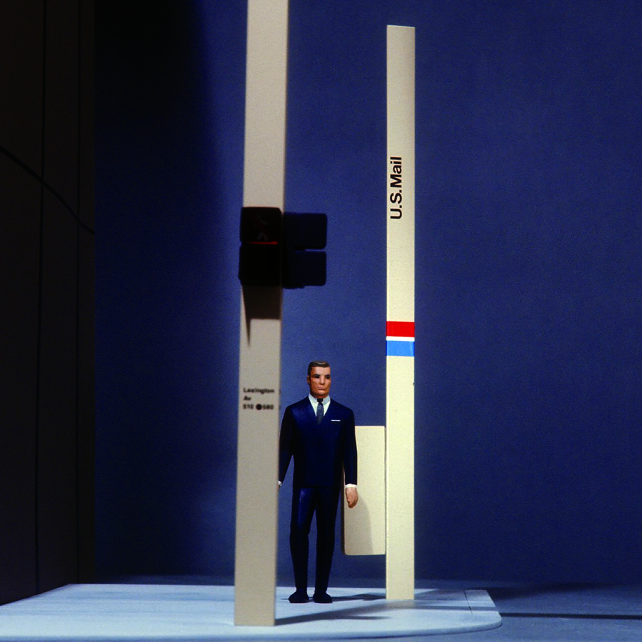 Miniature figure in a suit stands between tall, oversized U.S. Mail signposts against a blue background, creating a surreal, toy-like street setting.