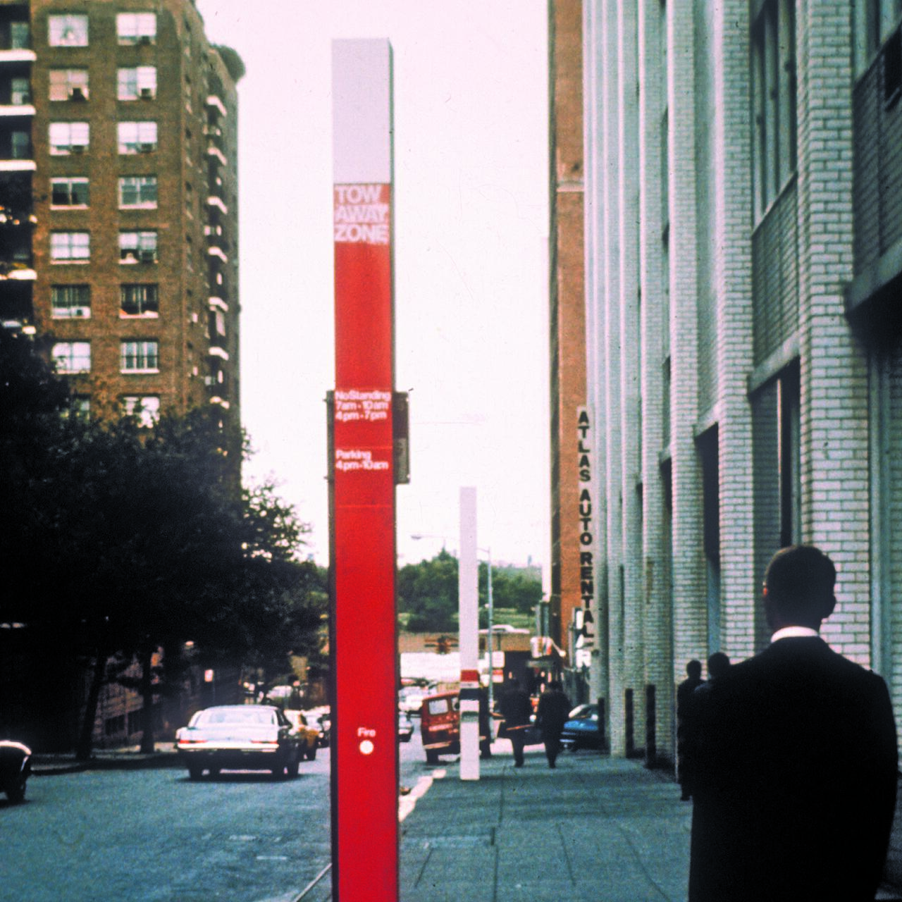 Street scene with a tall red and white "Tow Away Zone" pole, cars on the road, and pedestrians walking alongside buildings. A person in a suit stands on the right sidewalk near a brick building.