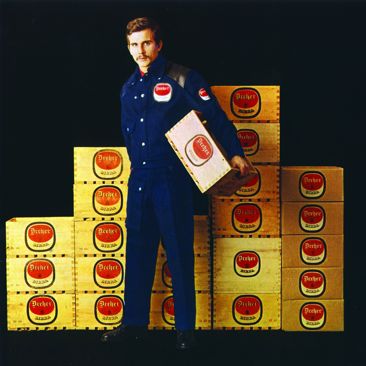 A man in a blue uniform holding a wooden crate stands in front of a stacked wall of similar crates labeled "Dreher" with a red logo, set against a dark background.