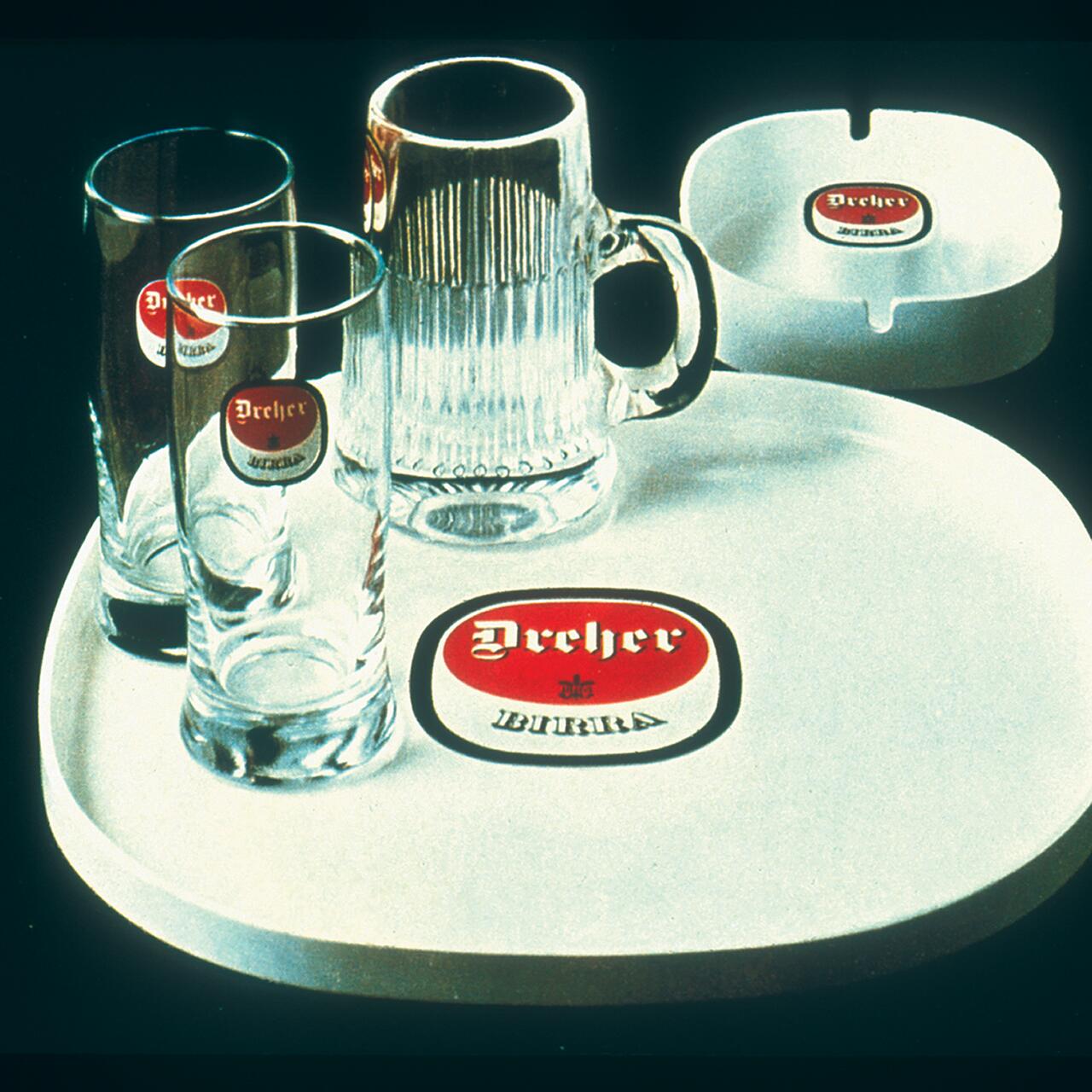 Beer-themed merchandise featuring two glasses and a mug with a red and white logo on a round tray, accompanied by an ashtray with the same logo.