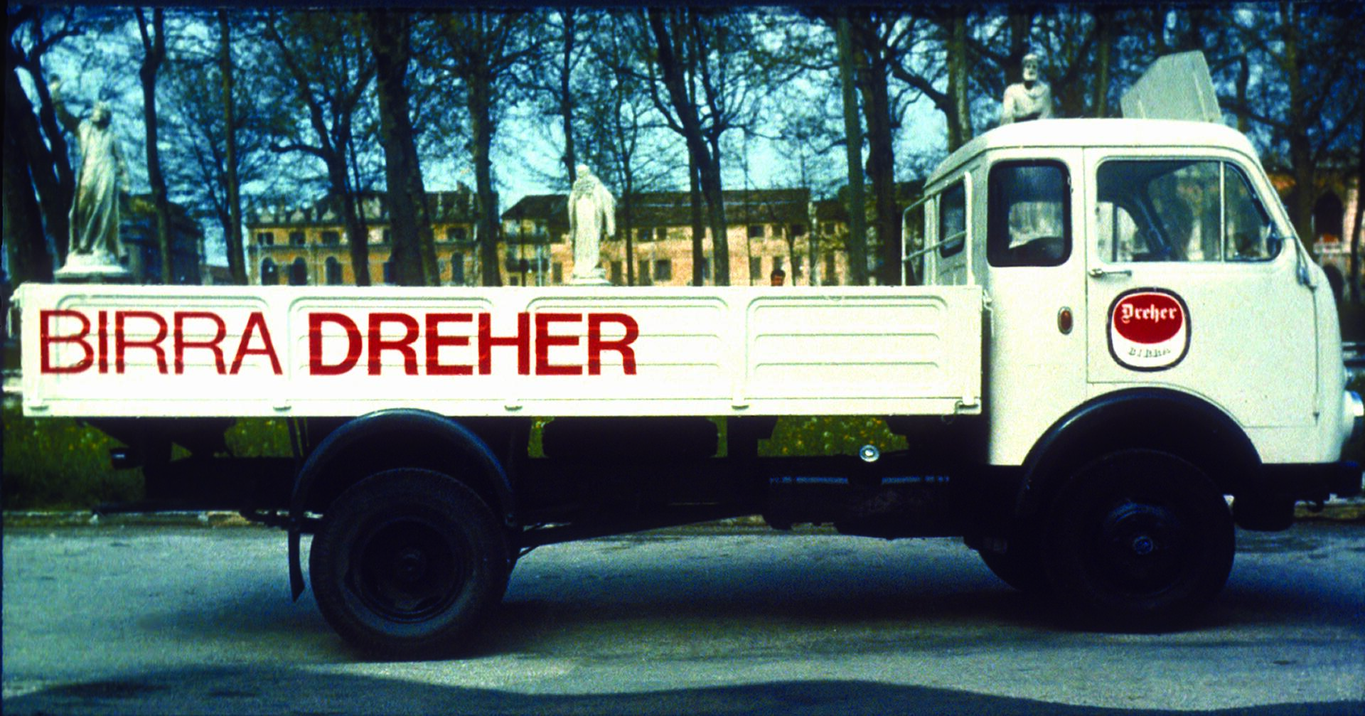 White delivery truck with "Birra Dreher" in red on the side, parked in a tree-lined area. Plaque with a similar design on the driver's door. Background features historic buildings partially visible through the trees.