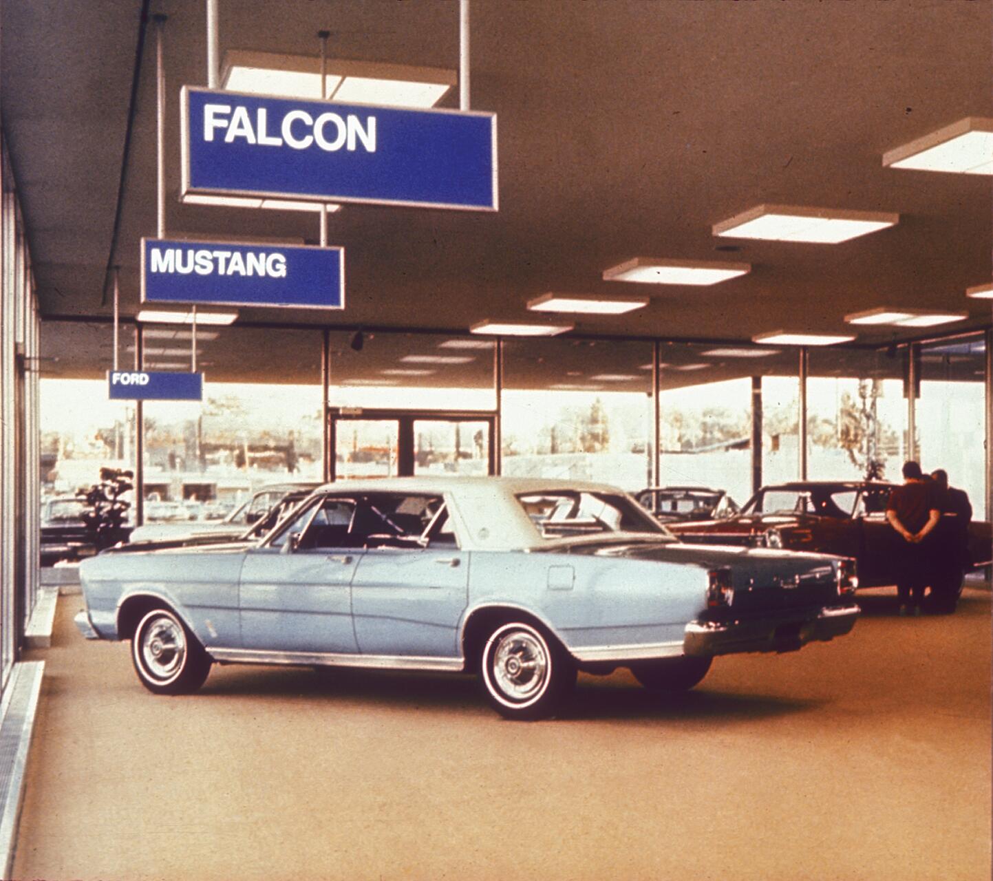 A vintage car showroom with a light blue classic car parked indoors. Ceiling signs display names of car models like Falcon and Mustang. Large windows in the background, with two people conversing near another vehicle.
