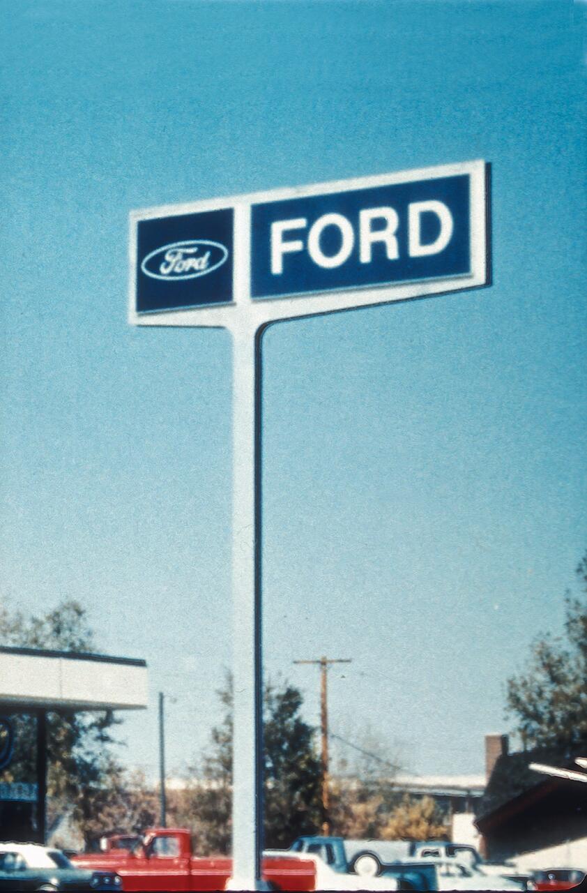 Ford dealership sign against a clear blue sky, with parked vehicles and trees in the background.