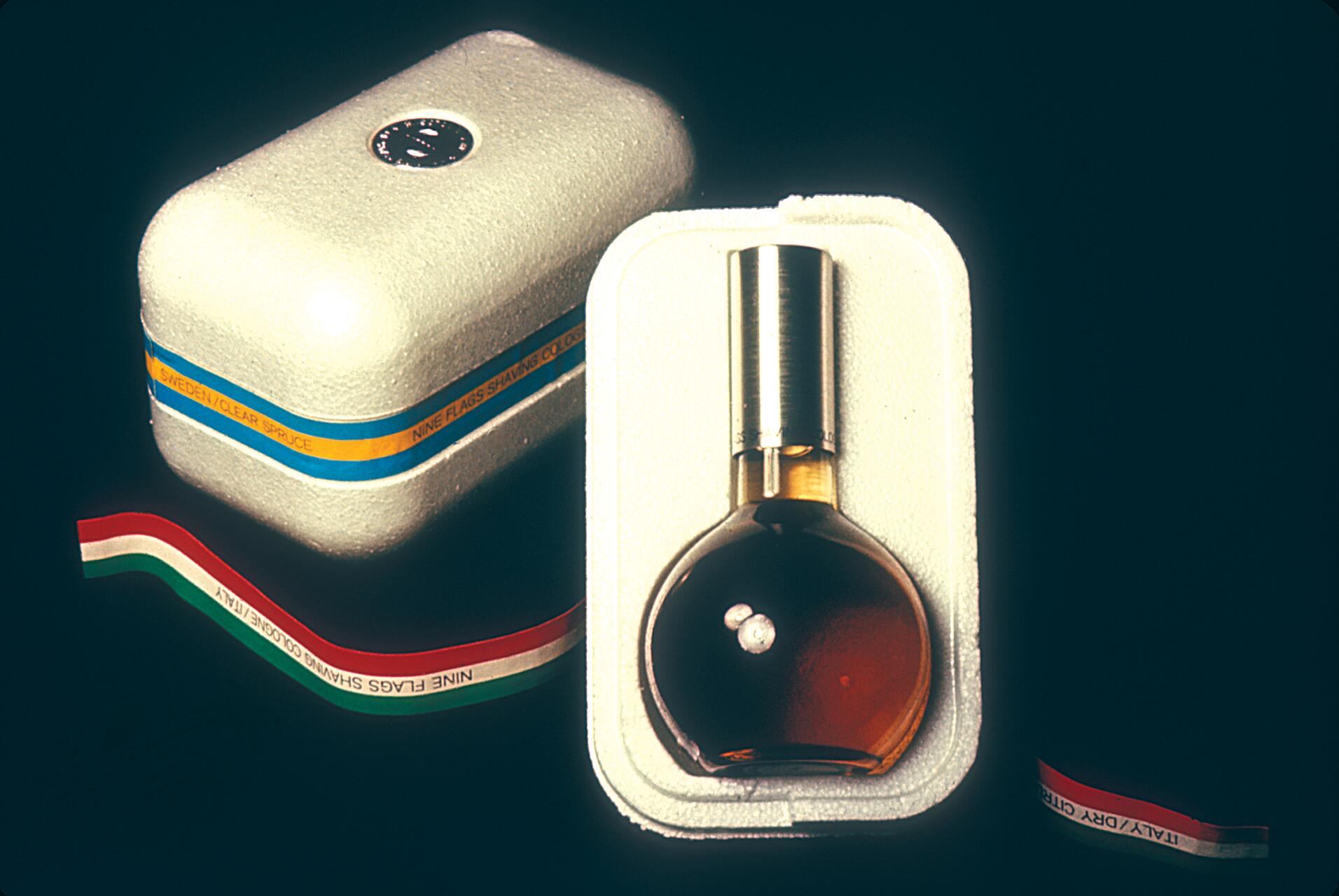 Cologne bottle with silver cap in a white foam container, featuring a striped box with text and tricolor ribbon.