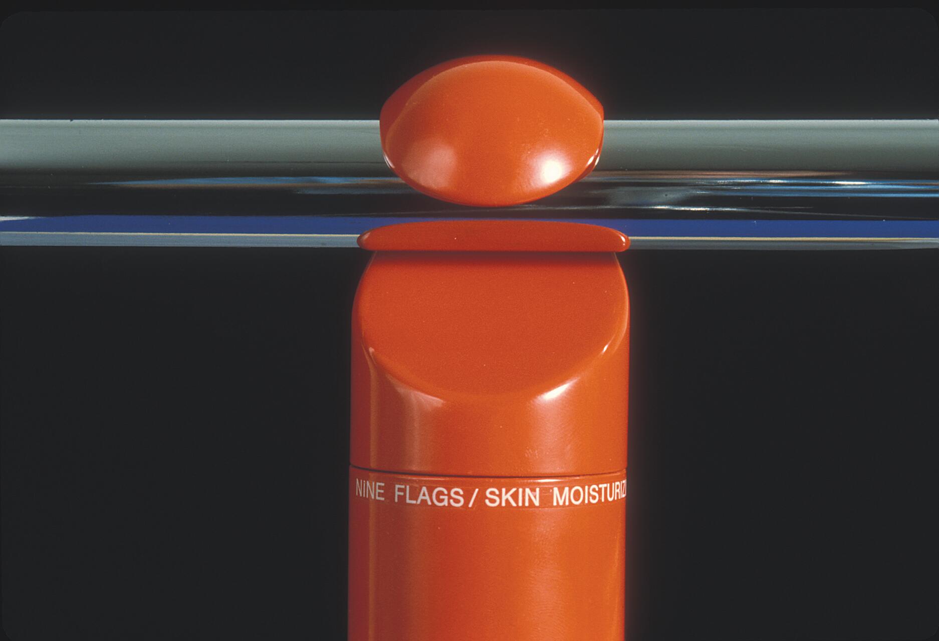 Orange cylindrical container with text "NINE FLAGS/SKIN MOISTURIZER" against a black background. A rounded cap balances above, and a horizontal chrome rod is visible in the background.