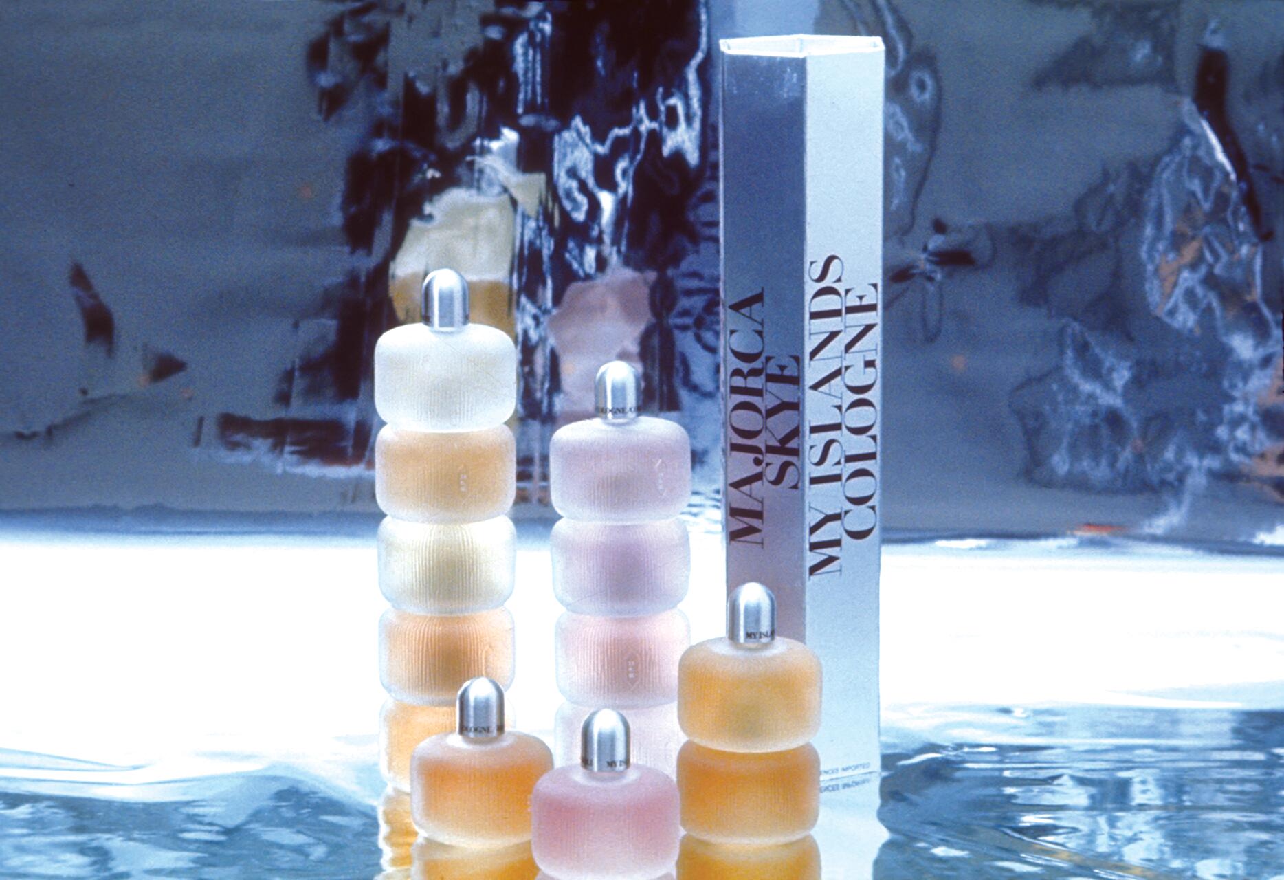 Stacked, three-tiered bottles with silver caps and pastel shades in front of a box labeled "Majorca Skye My Islands Cologne," on a reflective surface with a blurred background.
