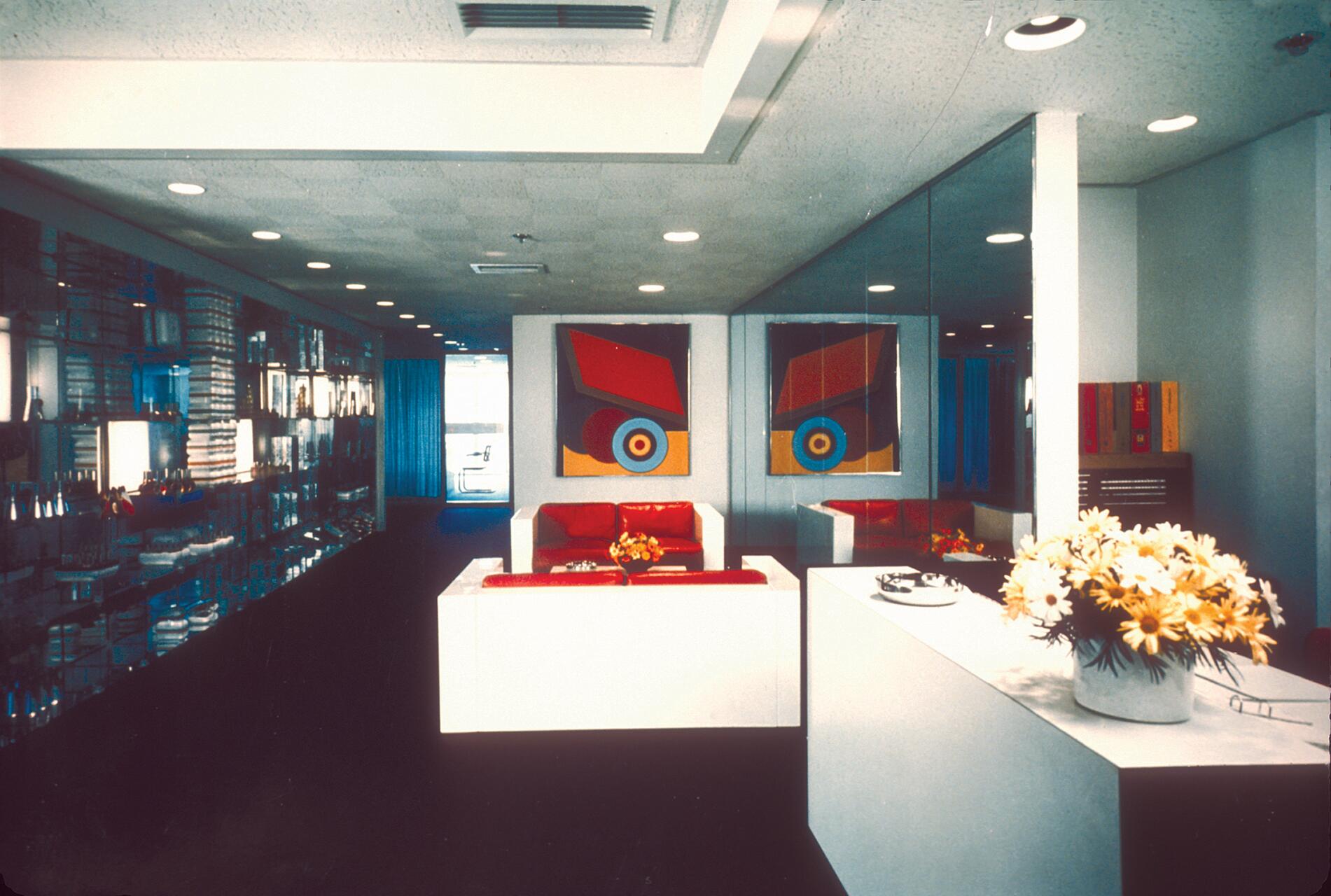 Modern lobby with mirrored walls, red seating, and abstract art. White counter with a vase of yellow flowers. Shelves on the left display items, and natural light filters through a window in the back.