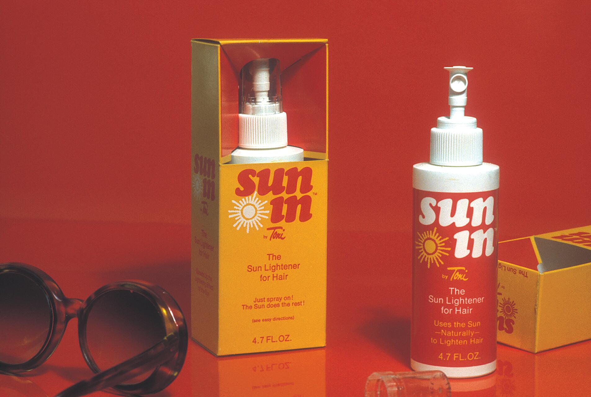 Hair lightener spray labeled "sun in" by Toni, with sun graphic, next to its yellow and red box on an orange background. Black round sunglasses lie beside the bottle.