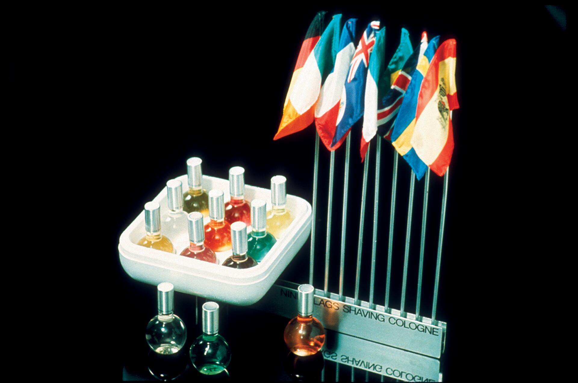 Various small bottles of colorful cologne next to a rack of miniature international flags, set against a black background.