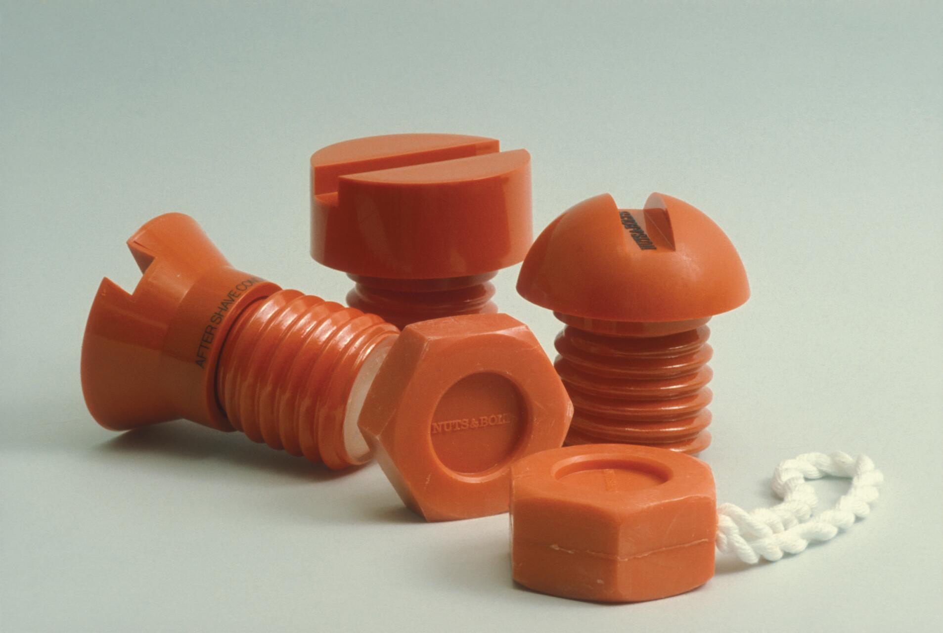 Orange plastic objects resembling oversized screws and bolts arranged together on a light background. A white rope is attached to one piece.