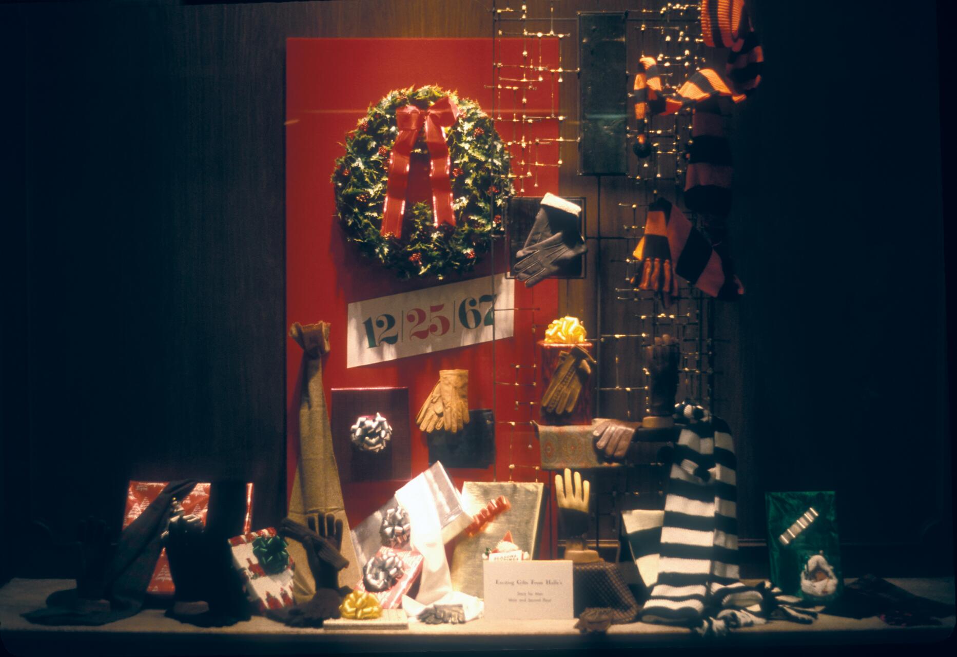Store window display with a festive wreath, gloves, scarves, and wrapped gifts against a red backdrop. Elegant holiday decorations suggest Christmas, with a large date reading "12/25/67."