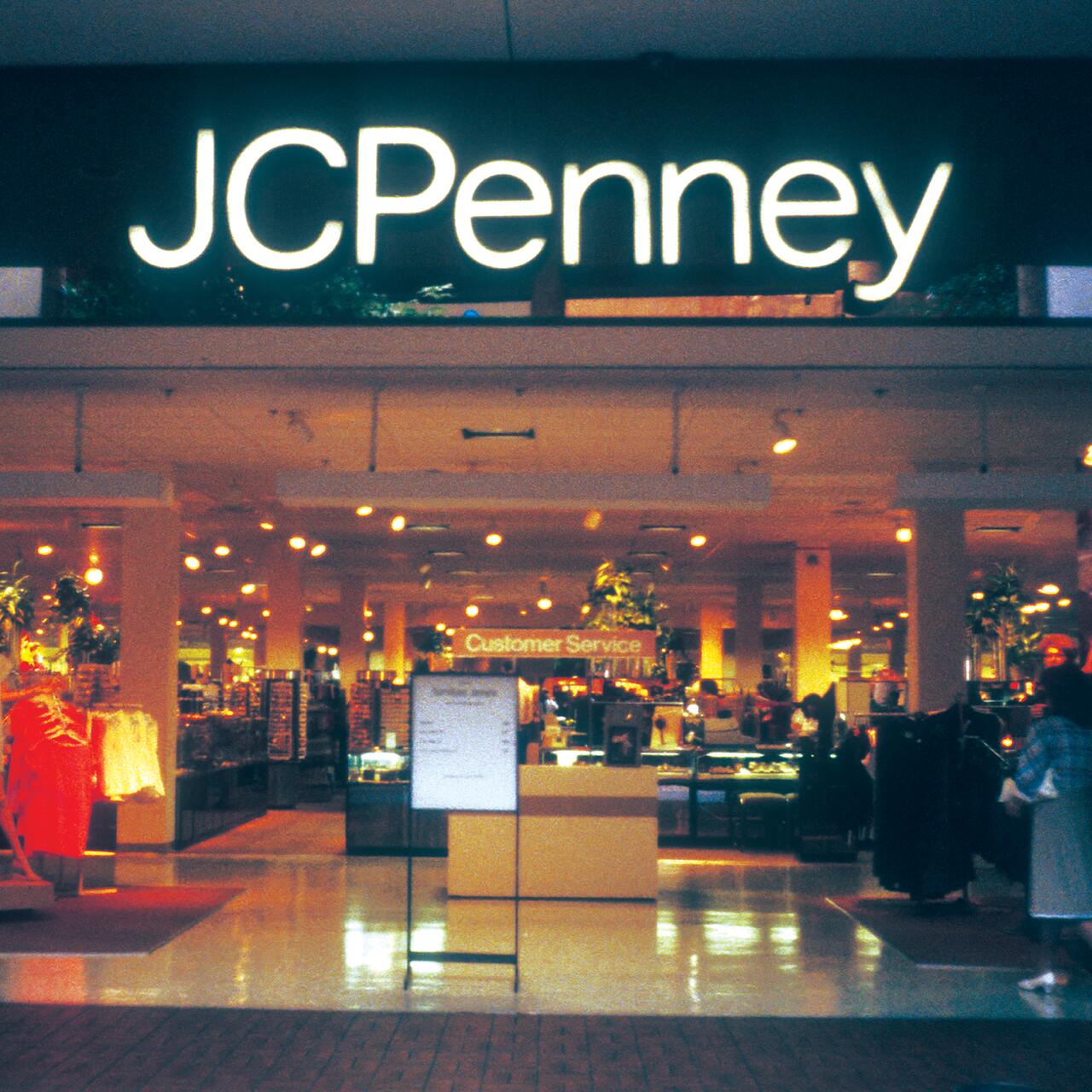 JCPenney store entrance with illuminated sign, featuring customer service desk and clothing displays inside.