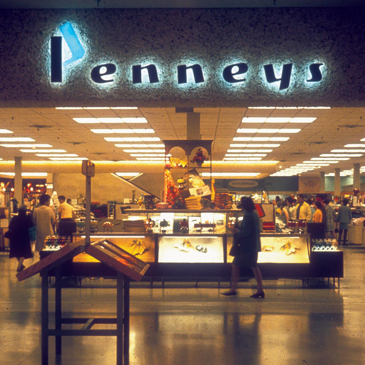 Vintage photograph of a Penneys department store interior, featuring people browsing various displays. The store sign is illuminated above, and the scene includes shelves stocked with colorful merchandise under warm, yellow lighting.