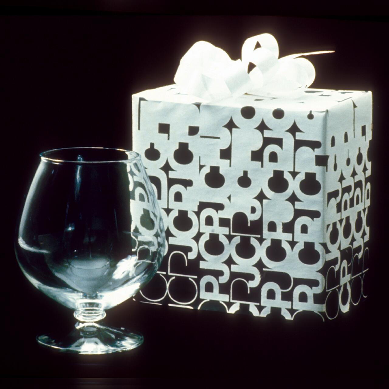 A clear snifter glass next to a white gift box with letter patterns and a large ribbon on top, set against a dark background.