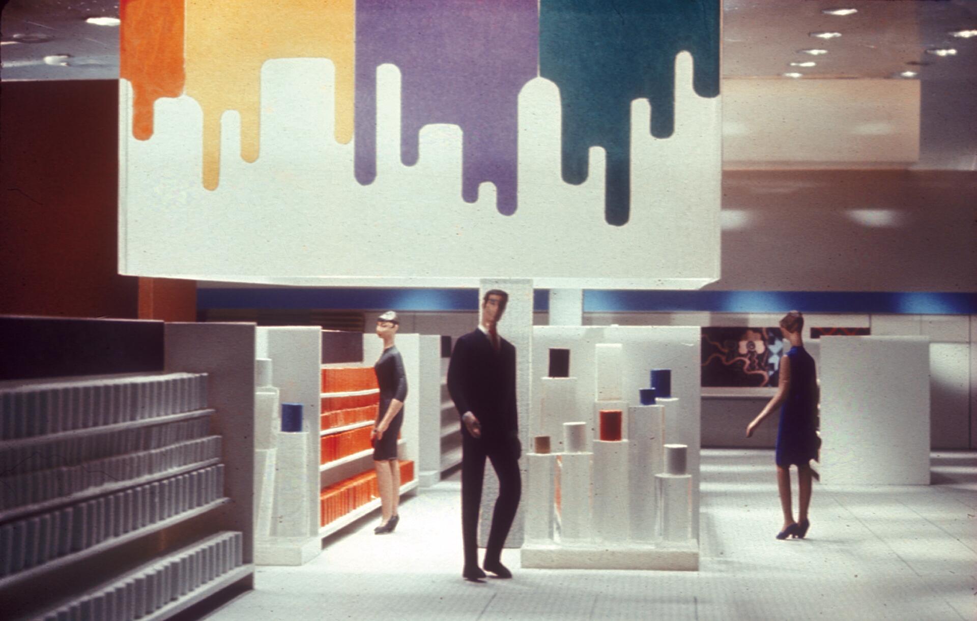A modern store interior with mannequins, white shelves, and colorful cylindrical displays. A white ceiling panel features dripping paint designs in orange, purple, and teal. Bright lighting enhances the contemporary atmosphere.