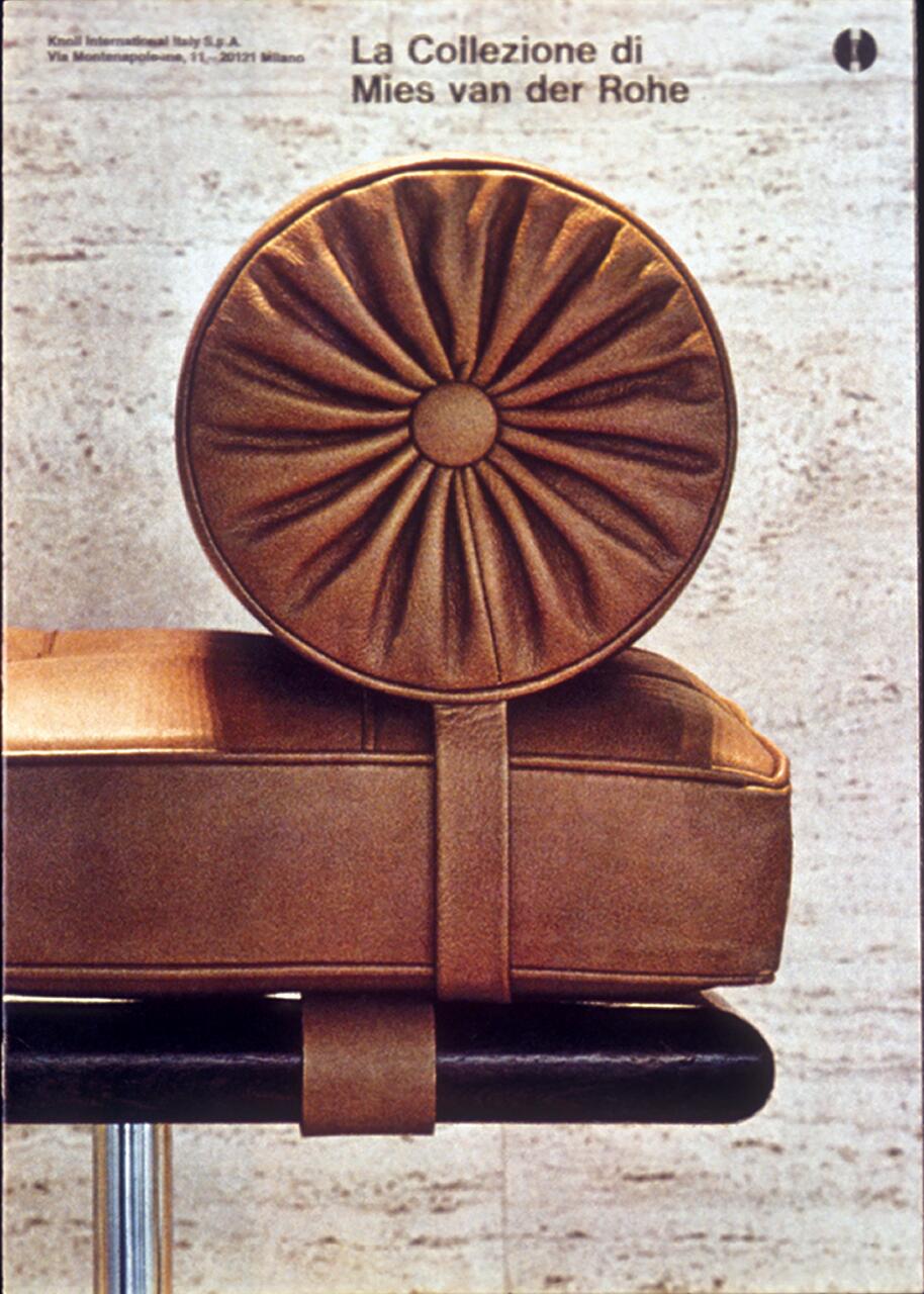 Close-up photograph of a tan leather cushion with a circular, tufted bolster, part of a Mies van der Rohe furniture design. The cushion is strapped to a black tubular frame against a textured stone background.