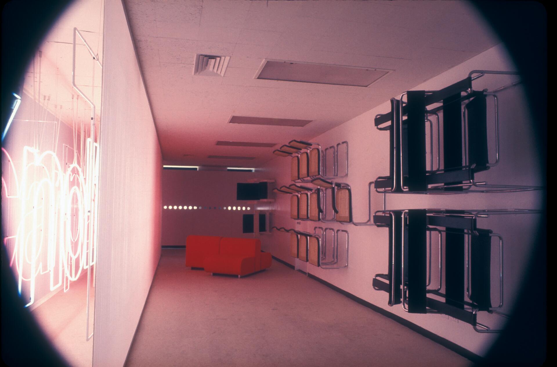 Dimly lit room with neon lights on the left wall, a red sofa in the center, and folding chairs stacked on the right wall. Warm, ambient lighting creates a retro-futuristic atmosphere.