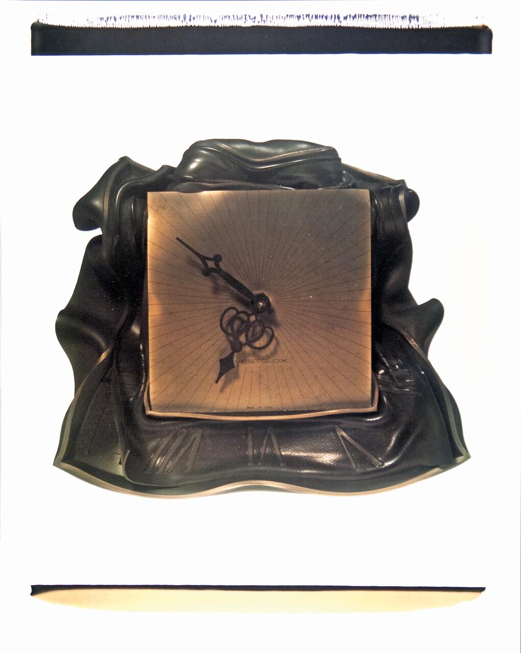 A square bronze sundial with ornate hands on a draped black fabric background.