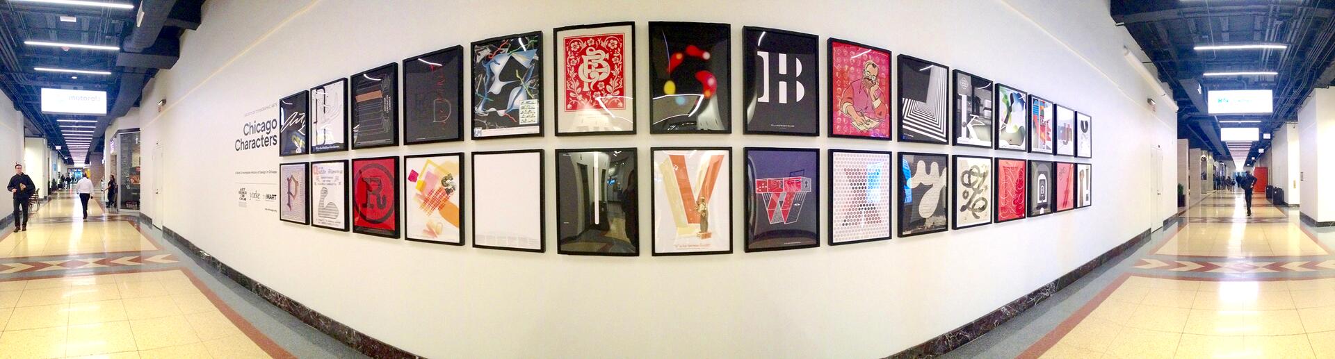 Framed art pieces hang on a long, white hallway wall, each featuring unique graphic designs. The hallway has a high ceiling with blue accents and is lit with bright overhead lights. People walk along the corridor.