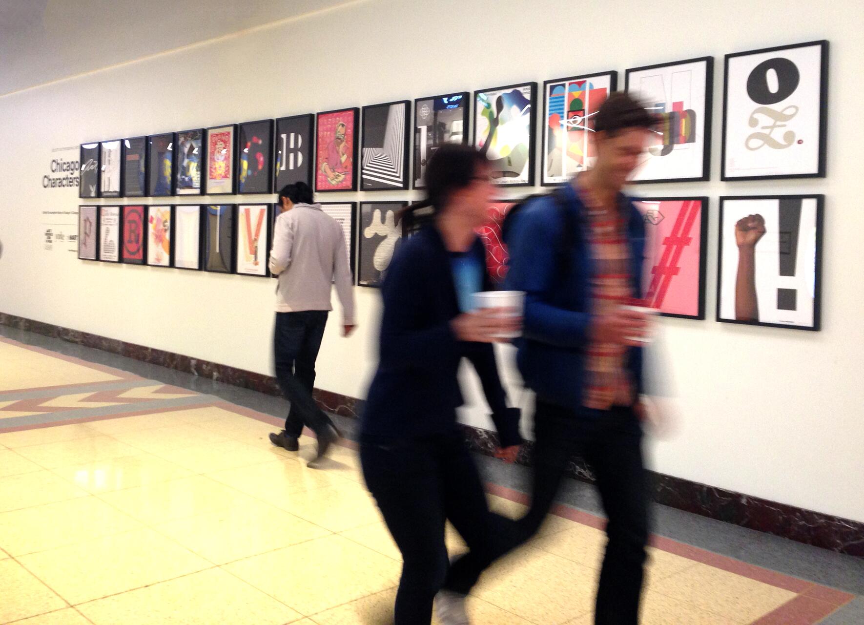 People walking past a gallery display of colorful framed artworks on a white wall, with one person closely examining the pieces.