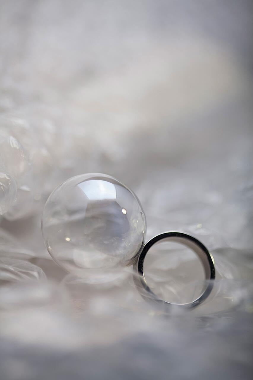 A clear glass sphere and a silver ring rest on a textured white surface, slightly out of focus, creating a soft, ethereal effect.