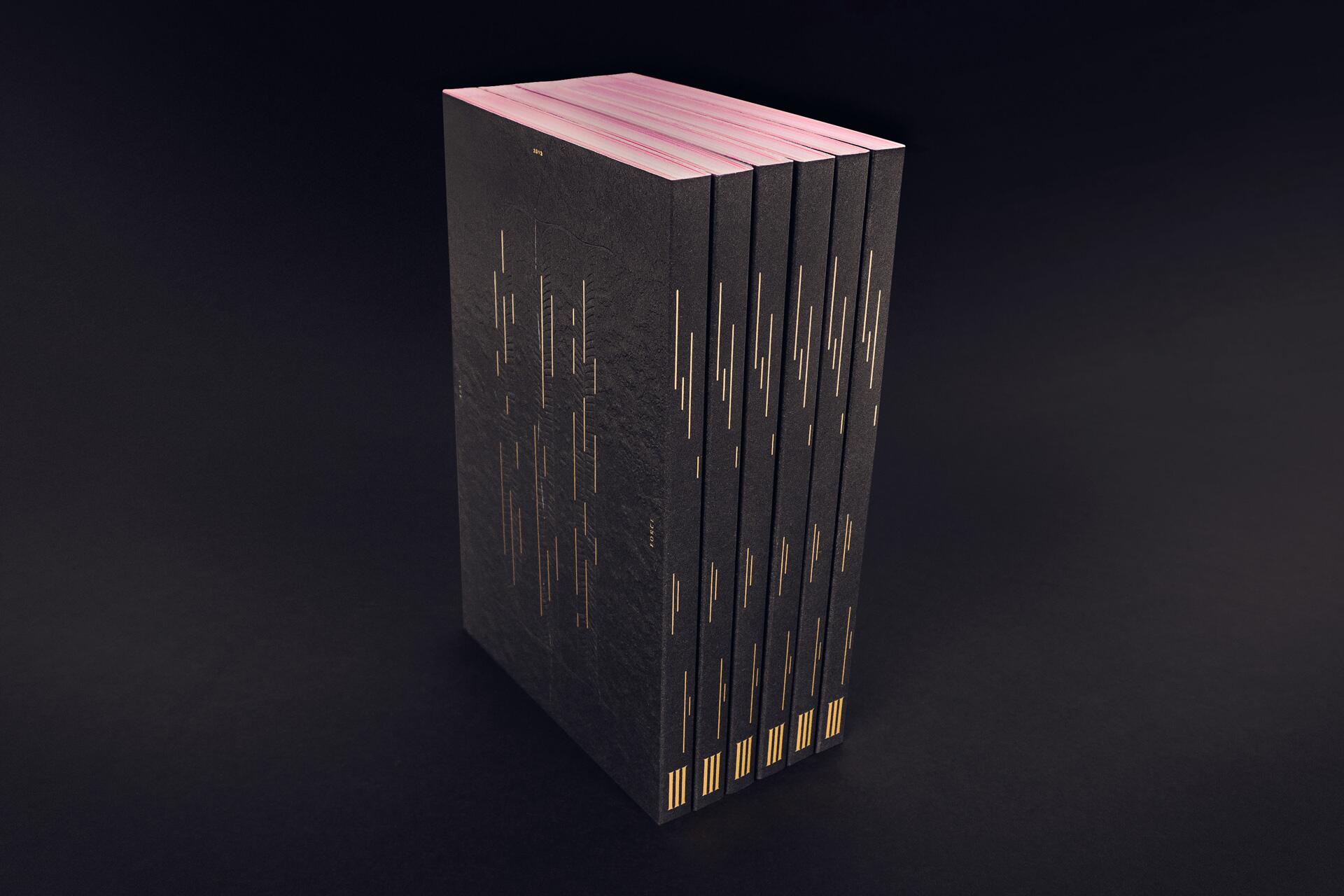 Stack of sleek black books with pink edges and gold line patterns on the covers, set against a dark background.