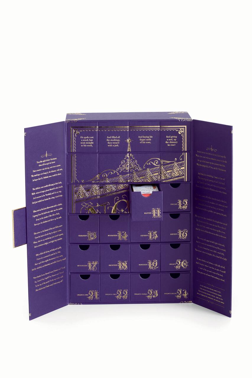 Purple advent calendar box with gold designs and numbered drawers. The box is open, revealing compartments for each day leading up to Christmas, with intricate decorative graphics and elegant script text on the inside panels.