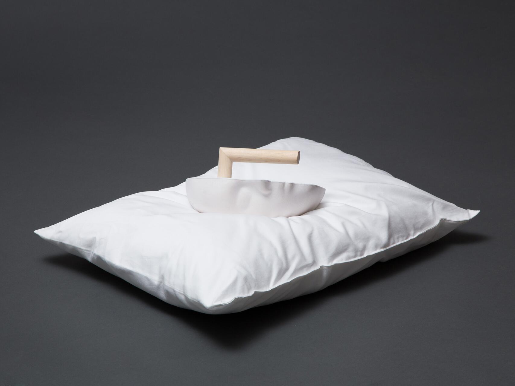 A white pillow with a minimalist gray vessel on top, featuring a light wooden handle, set against a dark gray background.