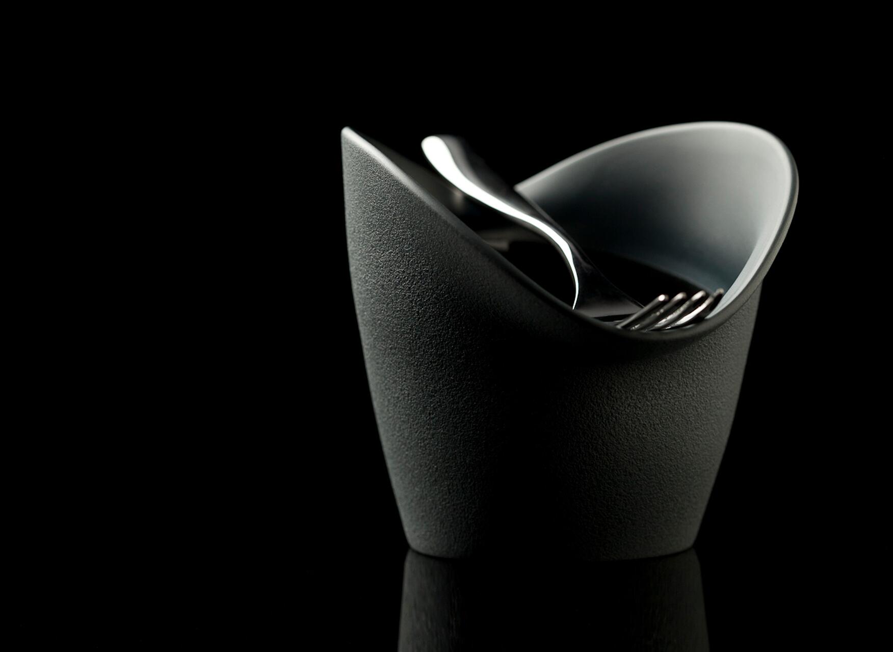 A sleek, modern gray bowl with an abstract design, holding a shiny silver fork and spoon, set against a black background.