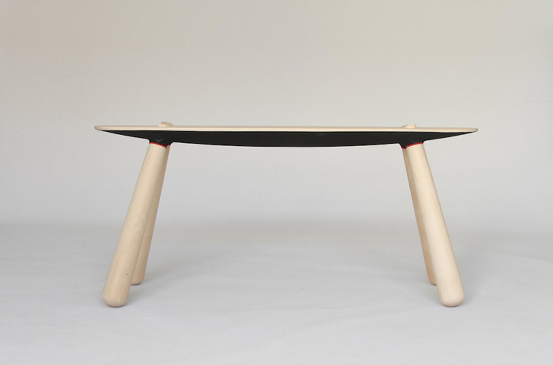 A modern table with a thin, rectangular top and four angled, cylindrical wooden legs. The table has a minimalist design with a black underside and red accents where the legs meet the tabletop.