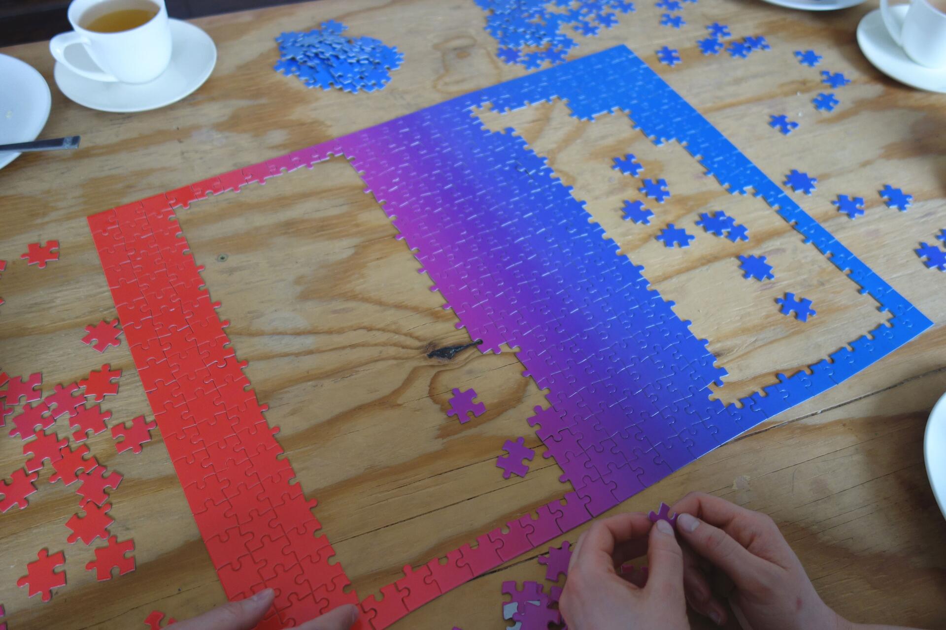 Color gradient puzzle being assembled on a wooden table, ranging from red to blue. Two hands are placing a piece. Several puzzle pieces and cups with saucers are scattered around.