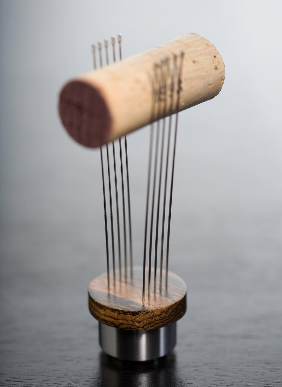 Wooden cylinder balanced on thin metal rods connected to a wooden and metal base, set against a dark surface.